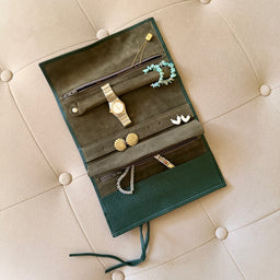 An open dark green leather jewelry organizer roll laid on a beige tufted leather chair, holding various jewelry items including earrings, a watch, and a bracelet.