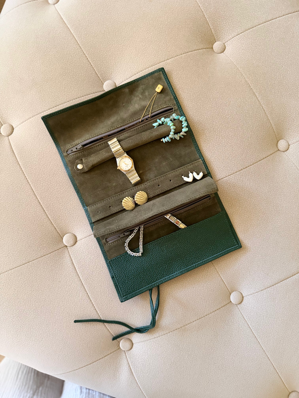 An open dark green leather jewelry organizer roll laid on a beige tufted leather chair, holding various jewelry items including earrings, a watch, and a bracelet.